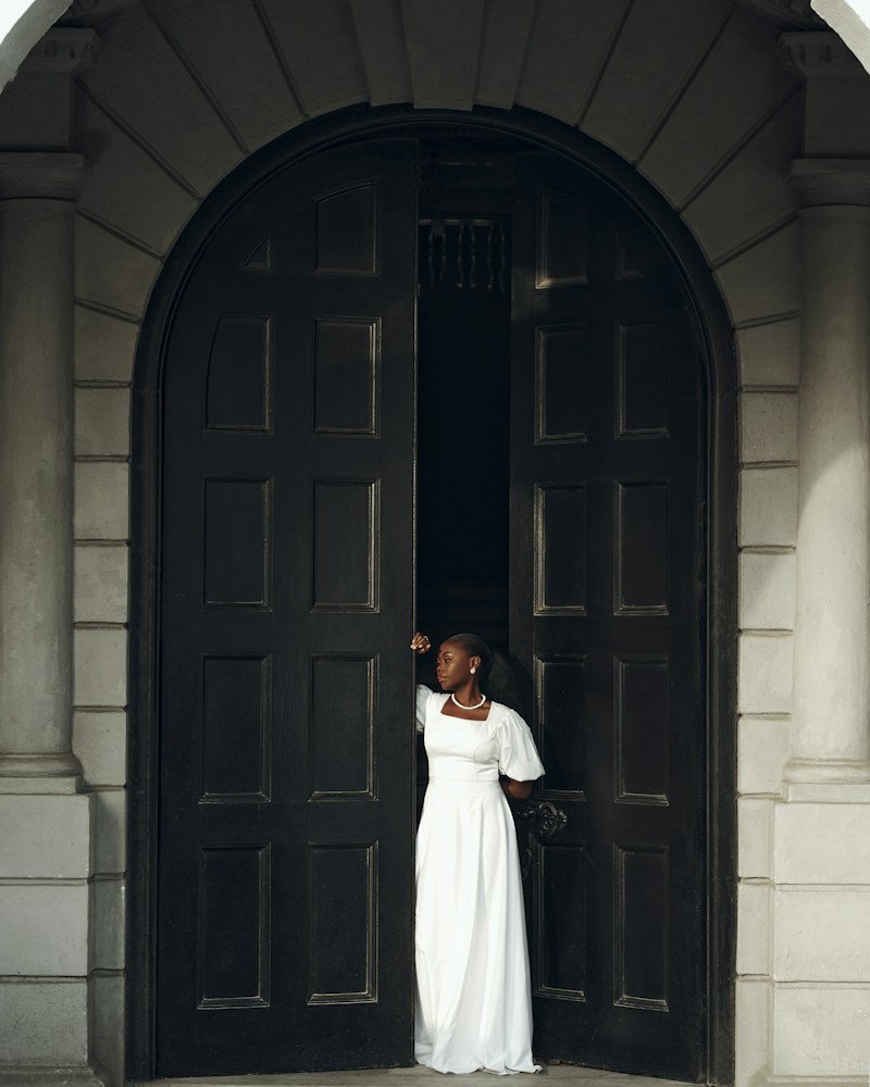 Editorial portrait of a woman in a white evening dress beside grand dark doors
